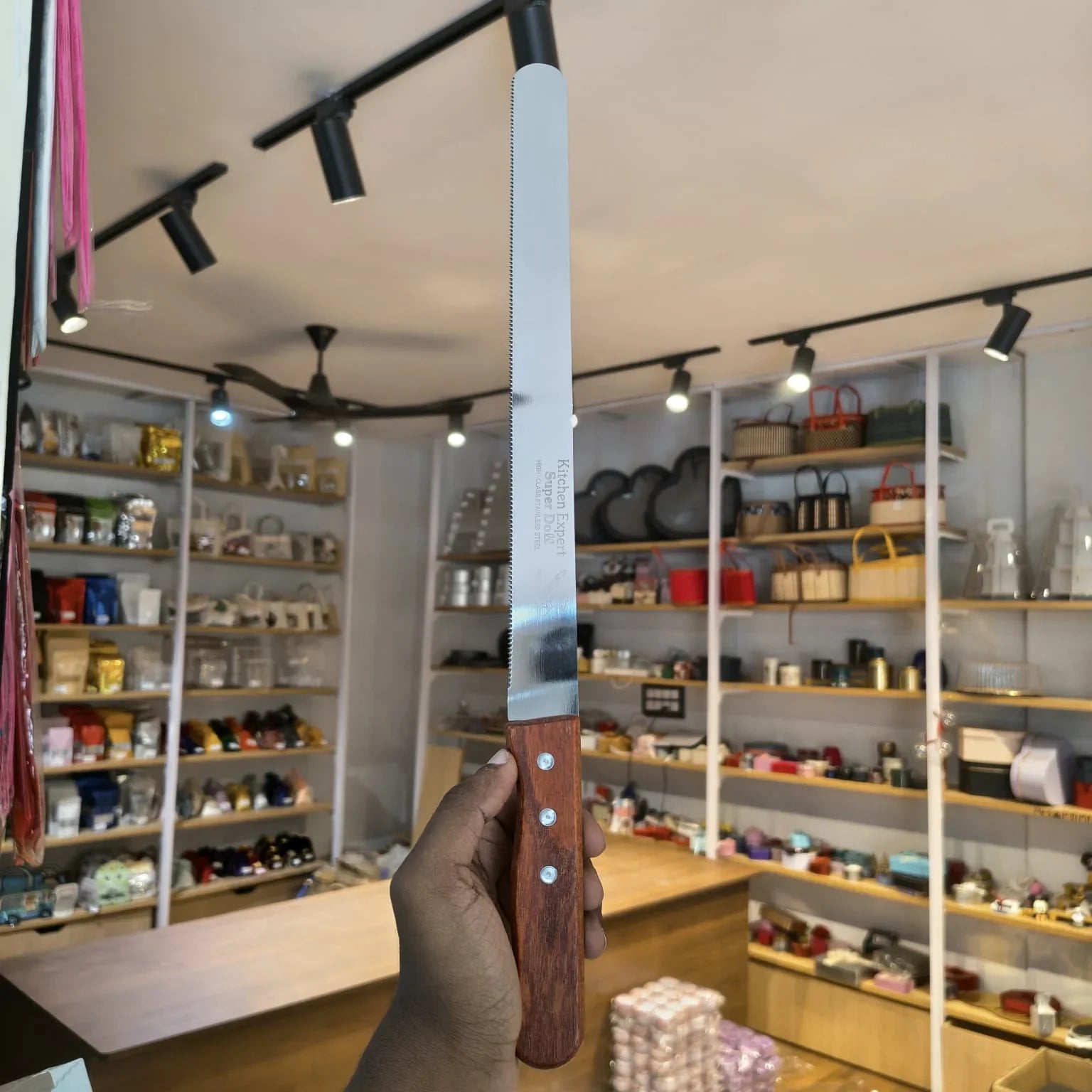 A hand holding a long serrated knife with a silver blade and a dark brown wooden handle inside a retail store with shelves of goods in the background.