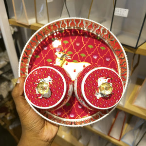A hand holds a round tray and matching cup set decorated with a festive red and gold pattern, featuring a white stag illustration and small gold clover accents.