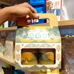 A hand holds a decorative gift box containing two small jars, photographed on a retail shelf. The packaging has a warm yellow and cream color scheme with traditional illustrated motifs and a cutout window showing the jars inside.