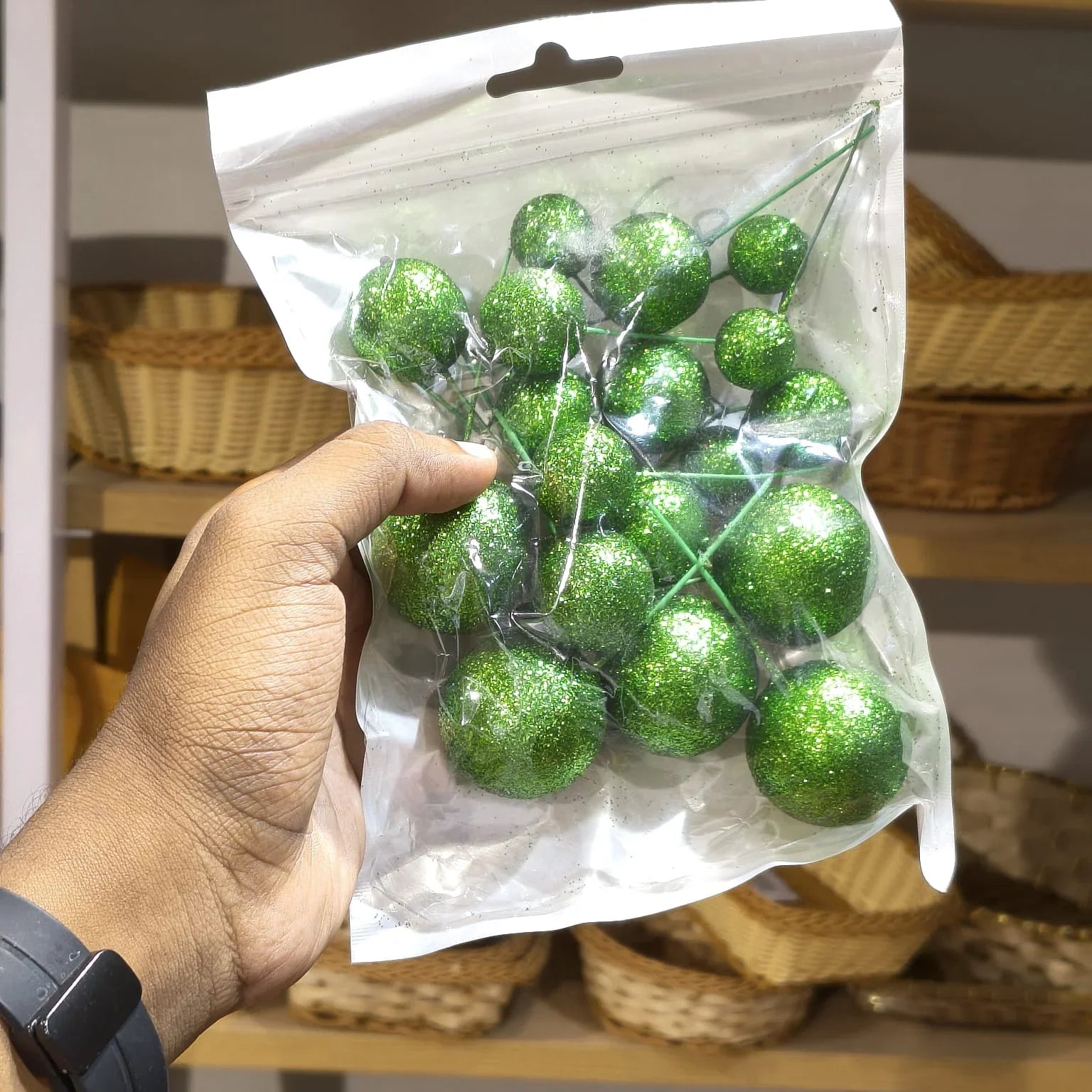 Hand holding a package of green glittery balls with wicker baskets in the background.