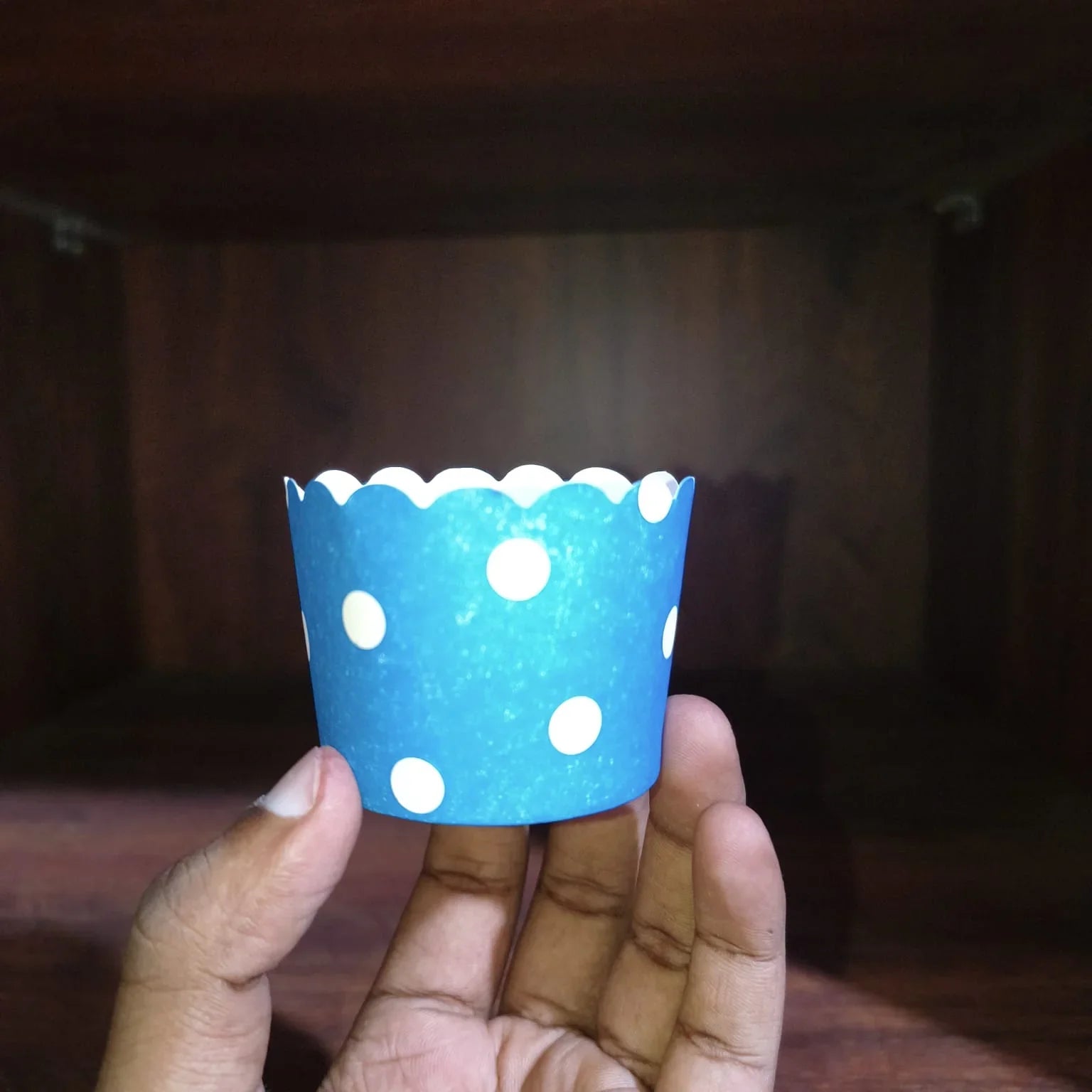 Hand holding a blue cupcake liner with white polka dots against a dark background