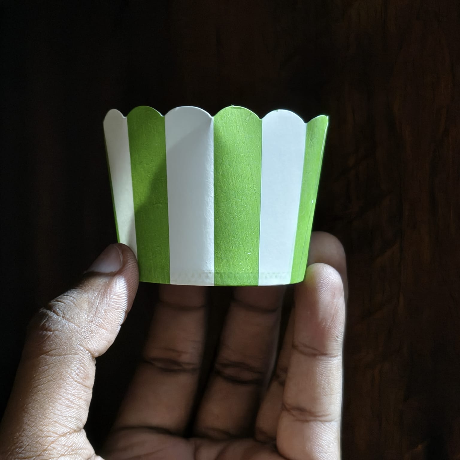 Hand holding a green and white striped paper cupcake liner against a dark background