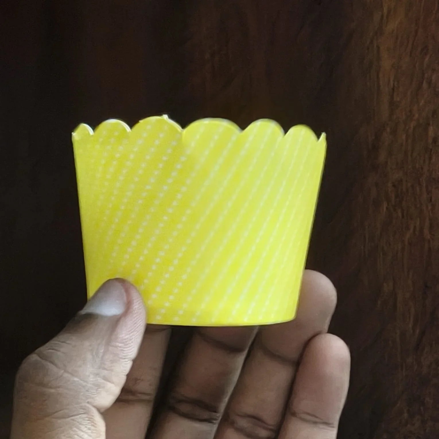 Hand holding a yellow scalloped-edge cupcake liner against a dark background