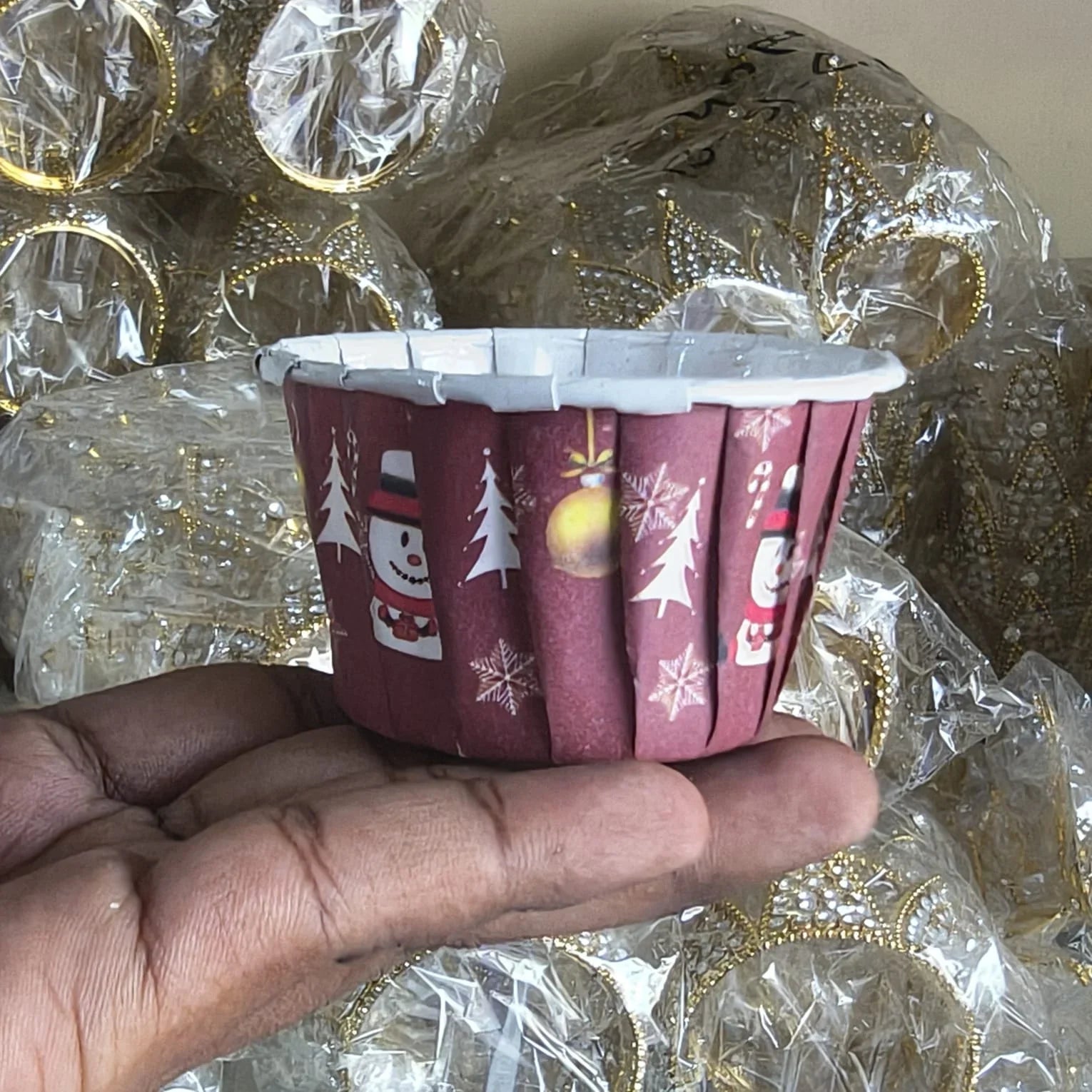 Hand holding a red Christmas-themed paper cup with decorative elements on a background of wrapped gold items.