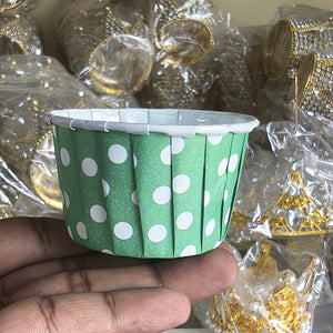 Green paper cup with white polka dots held by a hand against a blurred background of gold jewelry.