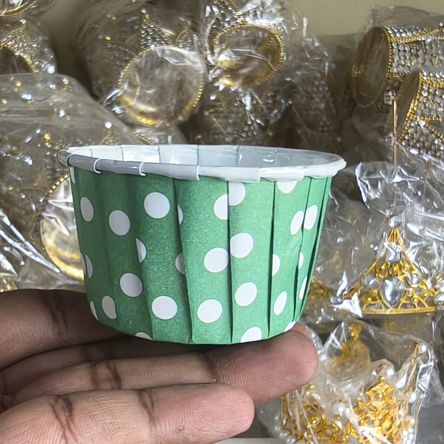 Green paper cup with white polka dots held by a hand against a blurred background of gold jewelry.