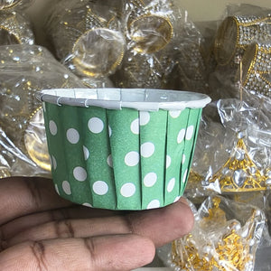 Green paper cup with white polka dots held by a hand against a blurred background of gold jewelry.
