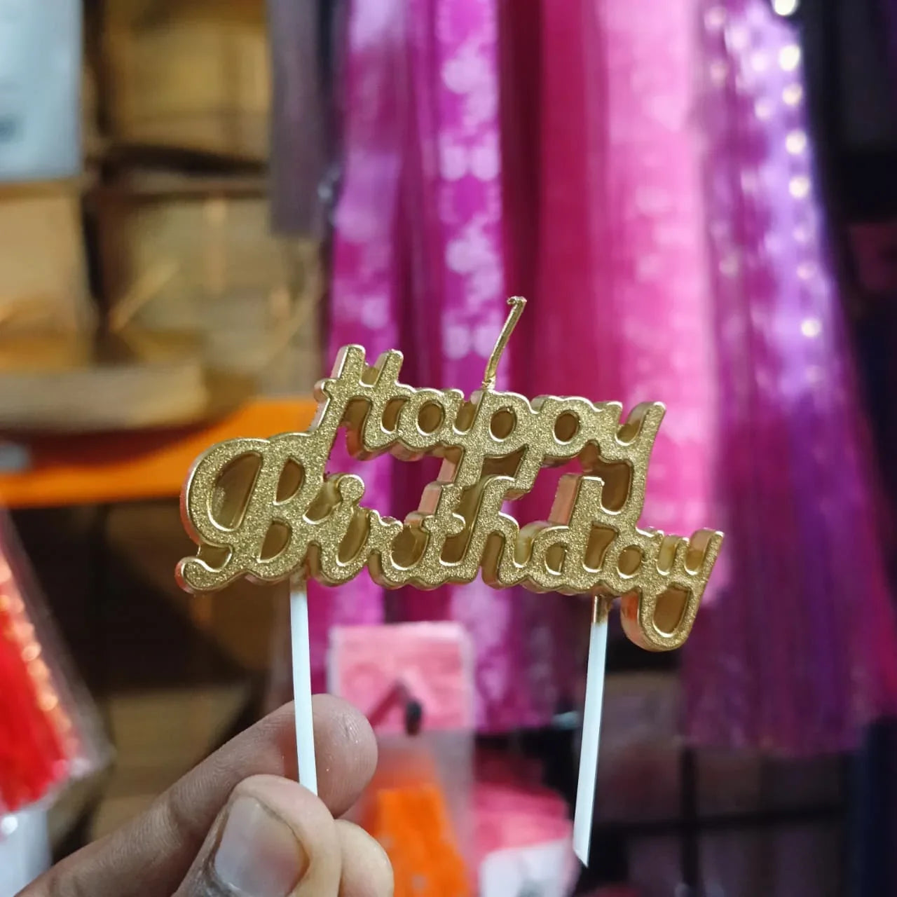Gold 'Happy Birthday' cake topper held in front of a colorful background with gift boxes.