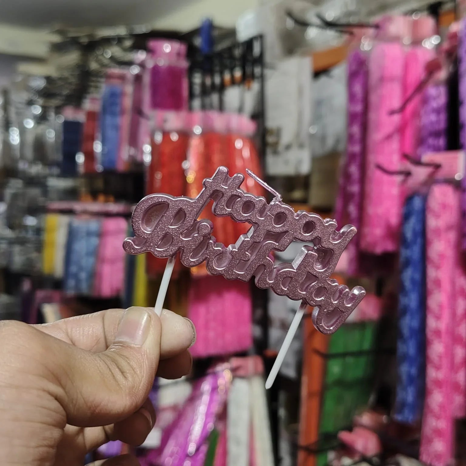 Hand holding a pink 'Happy Birthday' cake topper against a colorful store background.