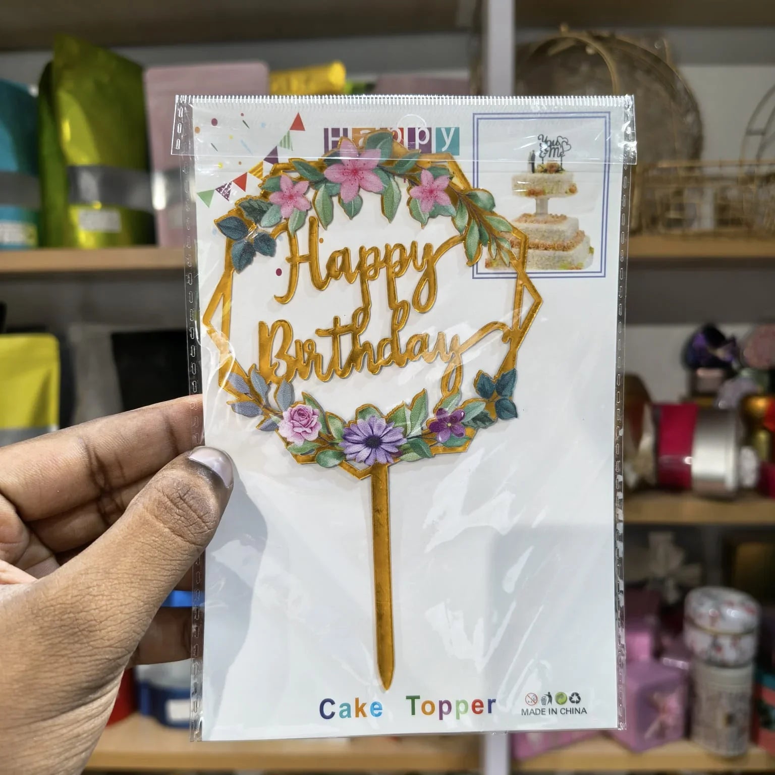 Hand holding a 'Happy Birthday' cake topper packaging with decorative elements.
