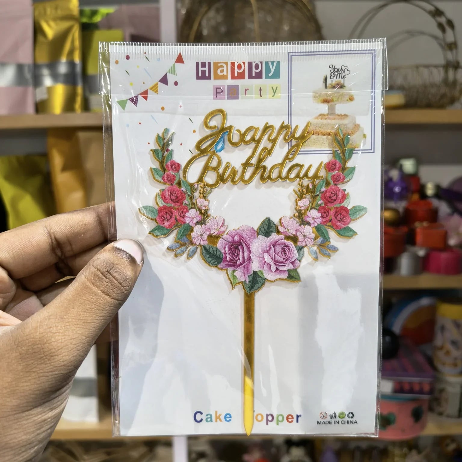 Hand holding a 'Happy Birthday' cake topper packaging with floral design.