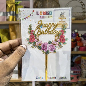 Hand holding a 'Happy Birthday' cake topper packaging with floral design.