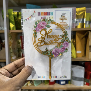 Hand holding a 'Happy Birthday' cake topper packaging with floral design.