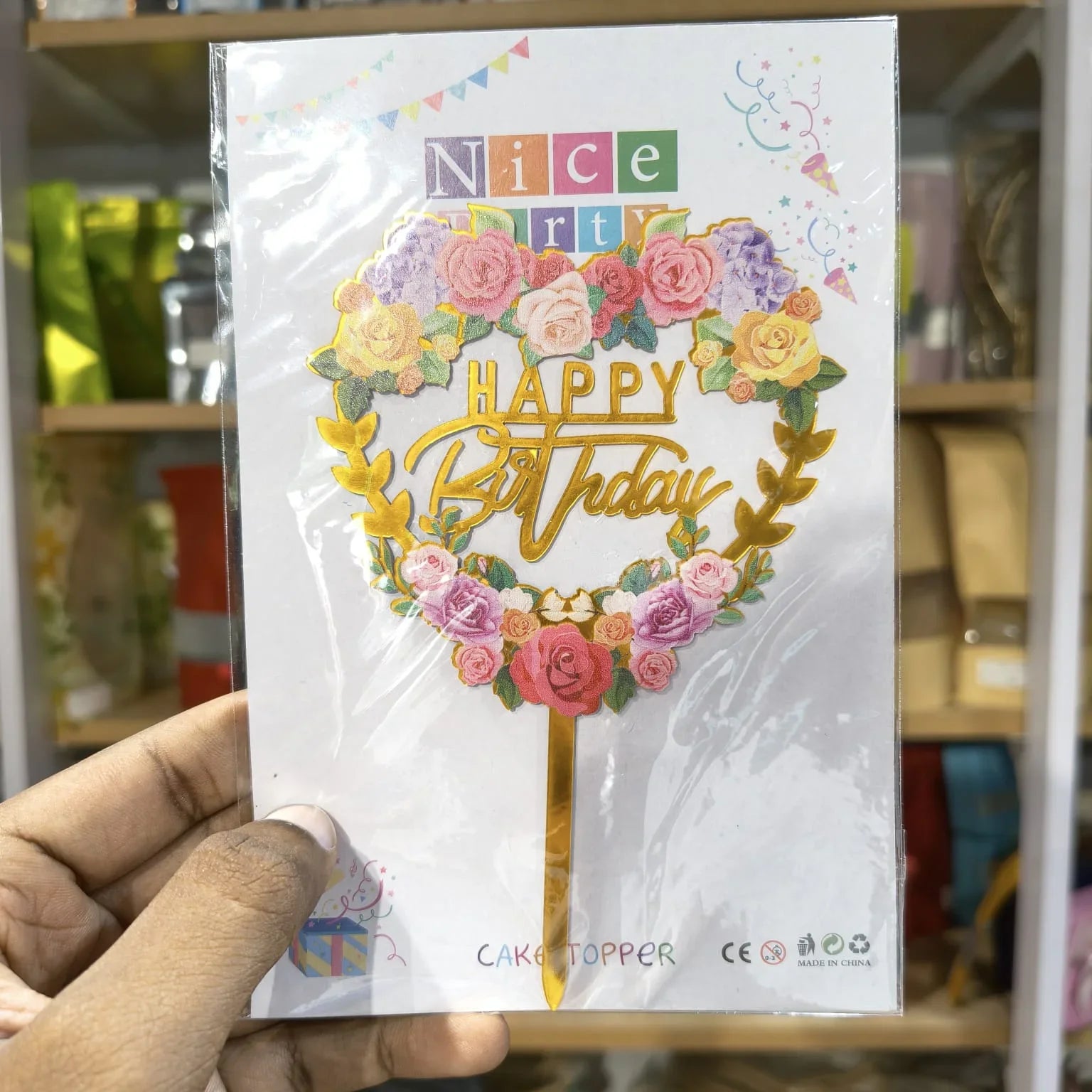 Hand holding a birthday cake topper packaging with floral design and text.