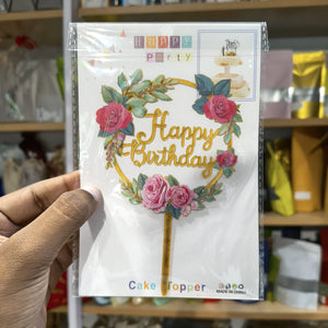 Packaged 'Happy Birthday' cake topper with floral design held by a hand.