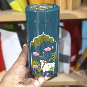 Teal cylindrical container with cow and floral design held by a hand against a blurred background