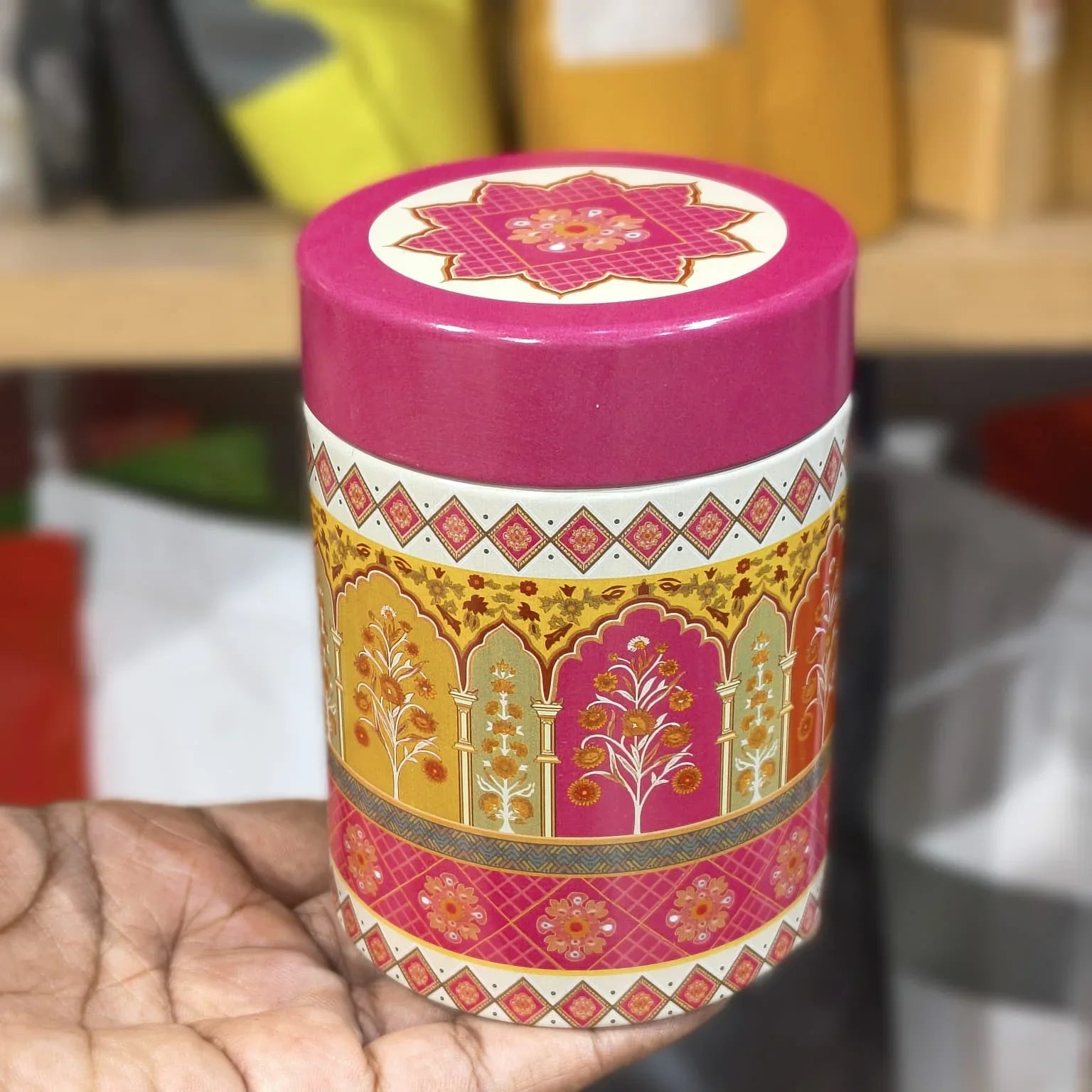 Decorative cylindrical container with pink lid and intricate patterns held by a hand.
