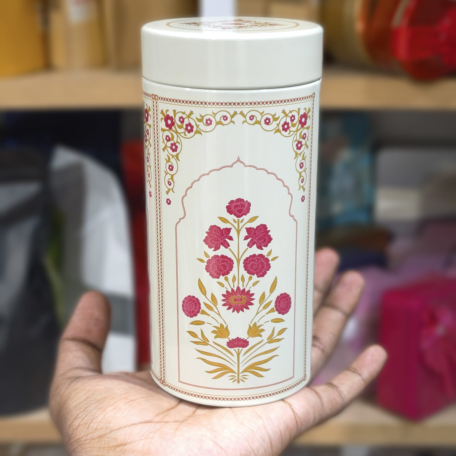 Hand holding a white cylindrical container with floral designs against a blurred indoor background