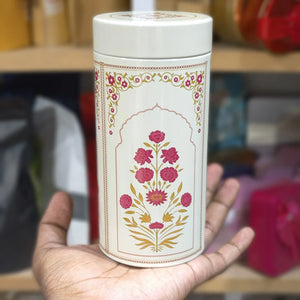 Hand holding a white cylindrical container with floral designs against a blurred indoor background