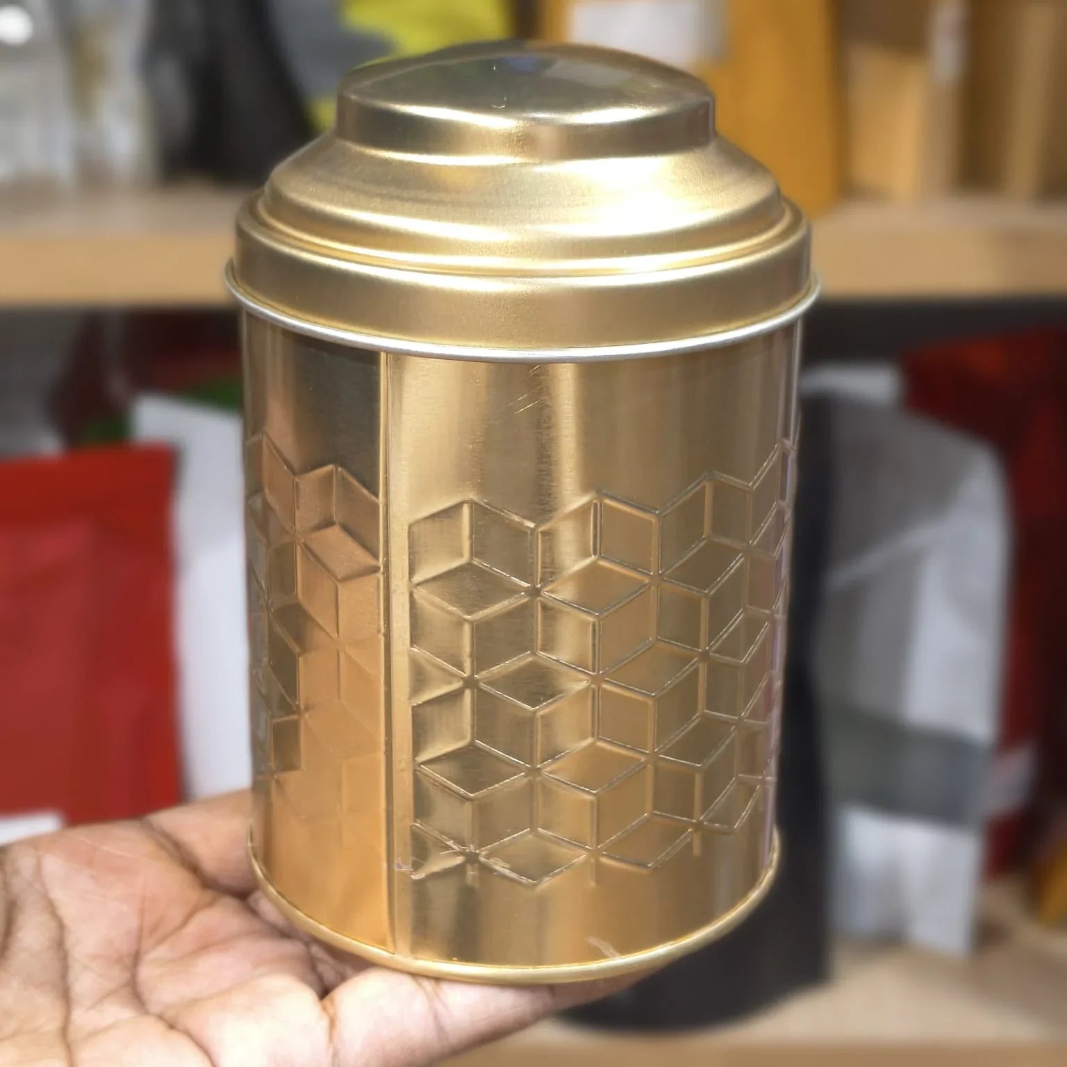 Gold cylindrical container with geometric pattern held by a hand against a blurred background