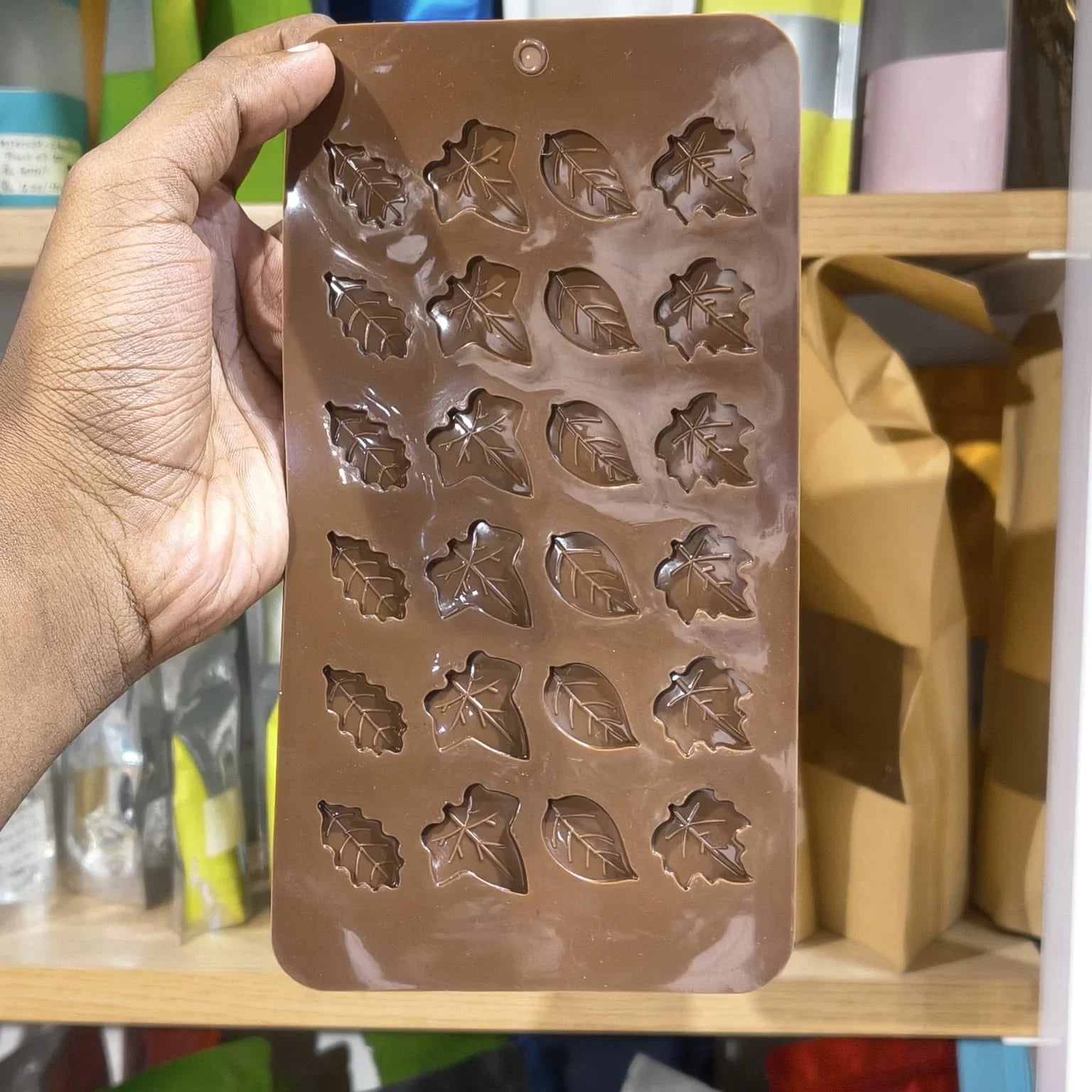 A hand holds a brown silicone mold with leaf-shaped cavities, shown against a background of shelves with various items and kraft paper bags.