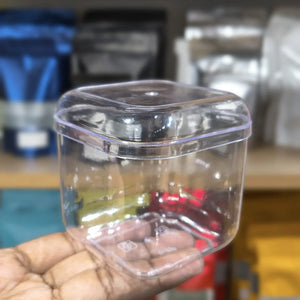 A clear plastic square container with a matching lid, held in a person's hand against a blurred background of shelves with packaged goods.