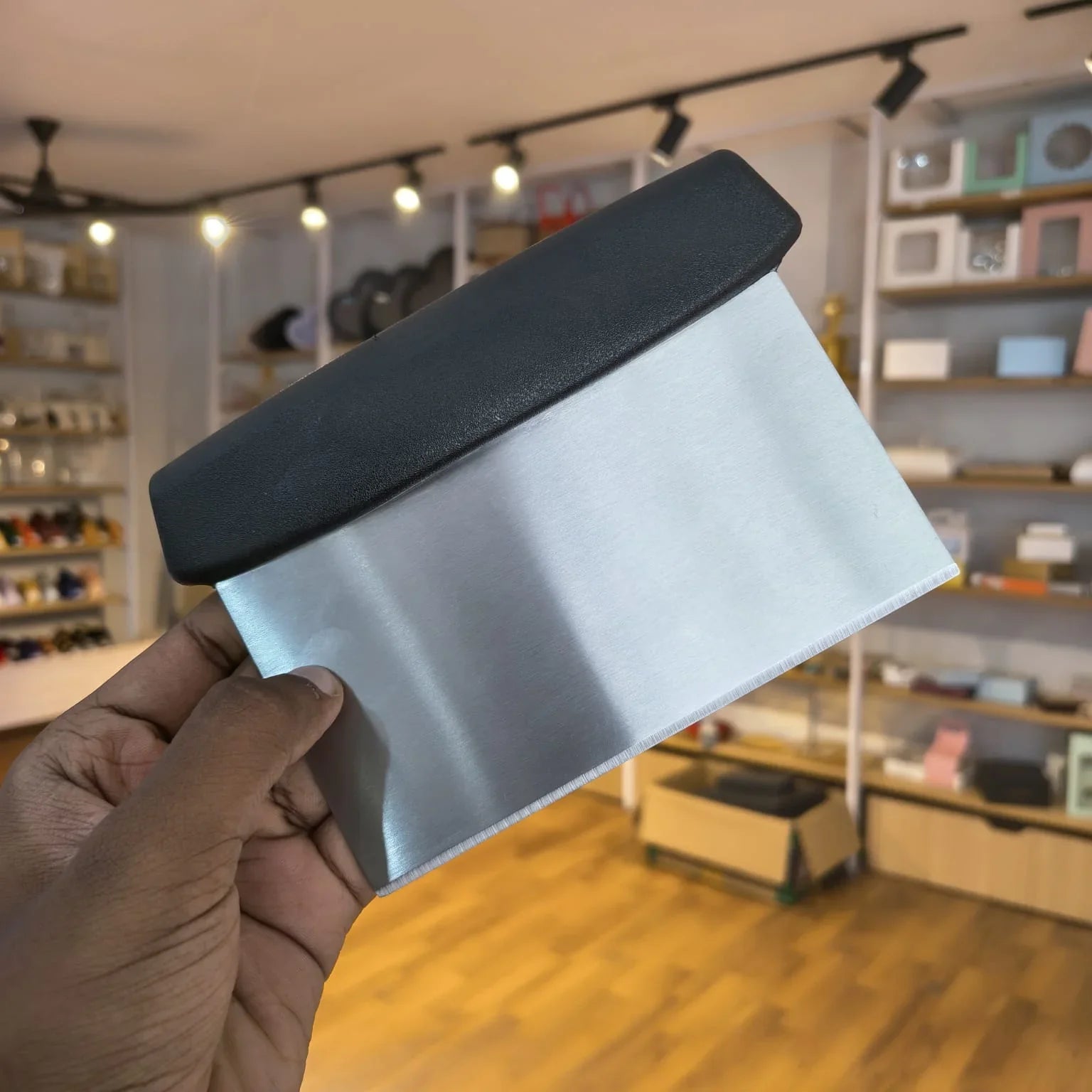 A hand holding a rectangular metal scraper or spatula with a brushed silver body and a black rubberized end, photographed in a retail store with shelves and fixtures blurred in the background.