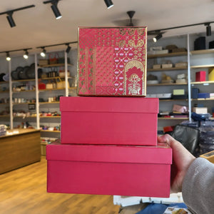 Three stacked gift boxes in a retail store setting, with the top box featuring a red and gold traditional patterned design and the bottom two boxes in solid bright pink. A hand is holding the stack on the right side.