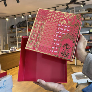 A person holds an open red gift box with a decorative red and gold patterned lid featuring floral and arch motifs, photographed in a retail store interior.