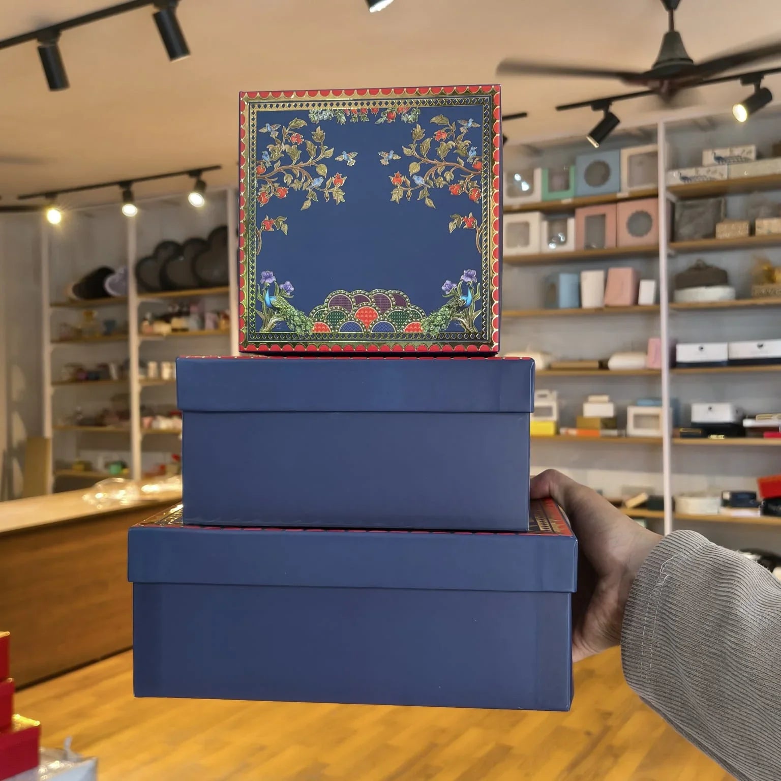 A person's hand holds a stack of three blue gift boxes in a retail store. The top box features a navy blue lid decorated with colorful floral embroidery and a red border.