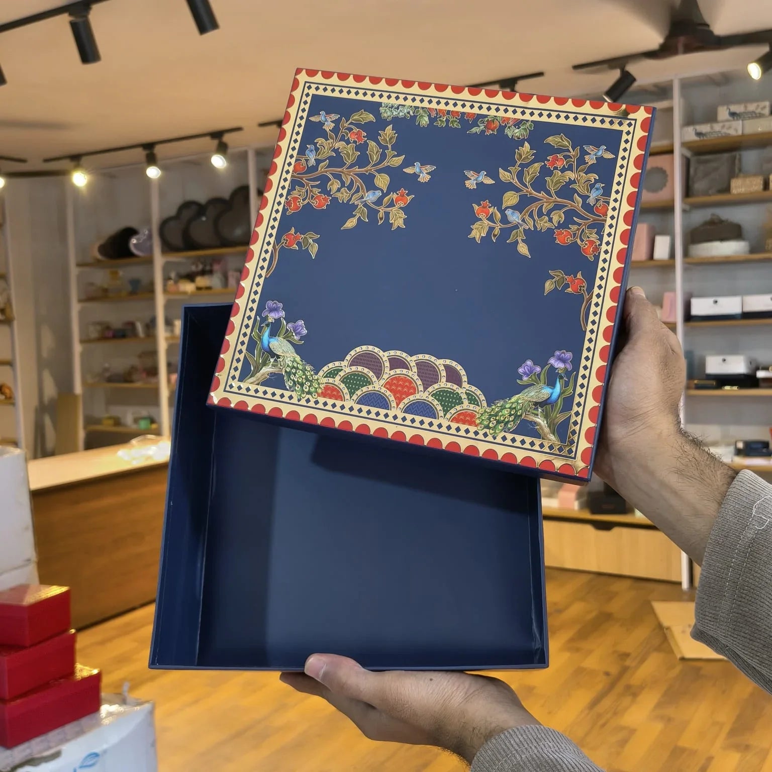 A navy blue decorative gift box with a floral and peacock pattern on the lid, held open by two hands in a retail store setting.