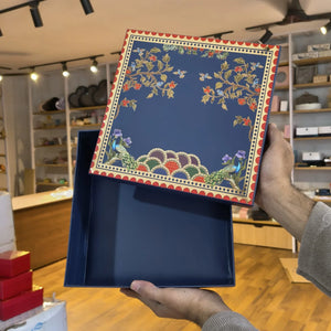 A navy blue decorative gift box with a floral and peacock pattern on the lid, held open by two hands in a retail store setting.