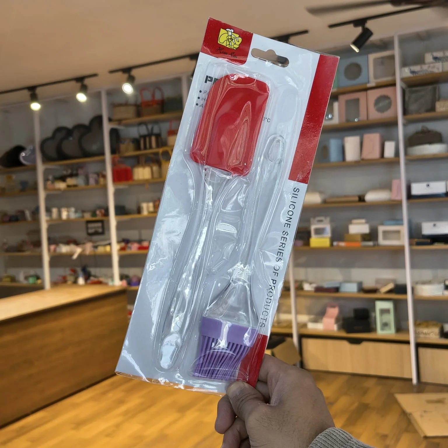A hand holds a packaged silicone cleaning brush set in a retail store. The product is in its original white and red blister packaging, showing a red-handled brush, a clear-handled brush, and a purple-handled brush.