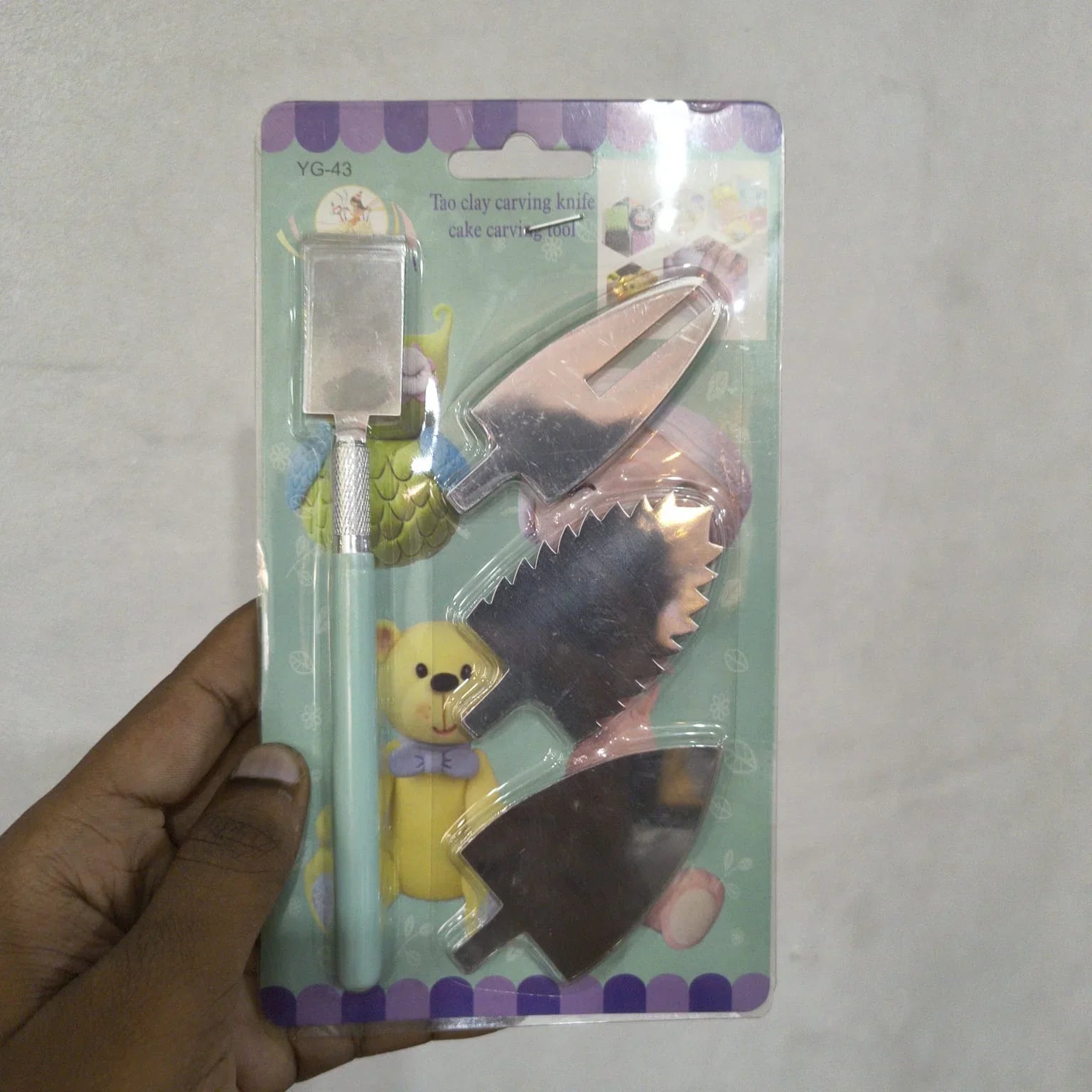 A hand holding a packaged set of three toy cake carving knives on a light gray background. The set includes a light blue knife with a silver blade, a pink knife with a silver serrated blade, and a dark brown knife with a silver blade.