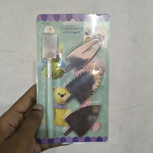 A hand holding a packaged set of three toy cake carving knives on a light gray background. The set includes a light blue knife with a silver blade, a pink knife with a silver serrated blade, and a dark brown knife with a silver blade.