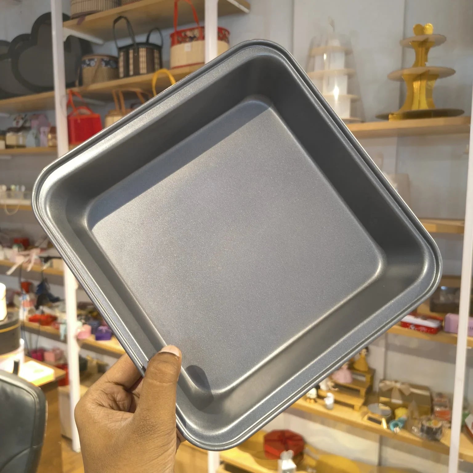 A hand holding a square, dark gray non-stick baking pan with raised corners, shown against a blurred background of a shop interior with shelves and items.