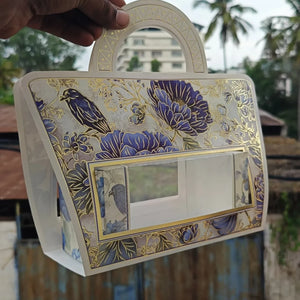 A hand holds a decorative handbag with a floral and bird pattern in blue, gold, and white, set against an outdoor background with palm trees and a building.
