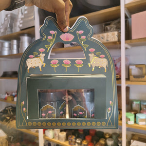 A hand holds a decorative teal gift box with gold and pink illustrations of cows, lotus flowers, and a central window displaying two small glass jars. The background shows a shop interior with shelves and jars.