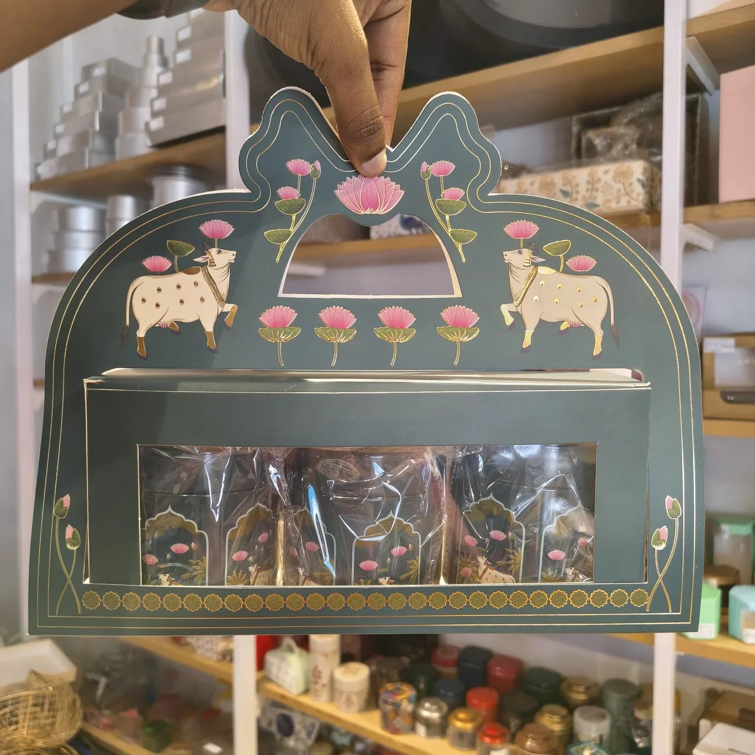A hand holds a decorative teal gift box with gold and pink hand-painted motifs, including two cows and lotus flowers, with clear plastic bags visible inside.