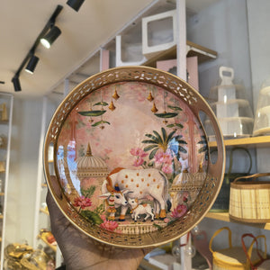 A hand holds a round decorative tray with a gold-toned cutout rim. The tray features a pastel pink background with a painted scene of a cow and calf, pink flowers, hanging bells, and domed architectural motifs.