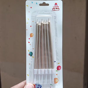 A hand holding a blister pack of six birthday cake candles. The candles are light brown with white tips and are packaged in a clear plastic case on a white backing card decorated with colorful confetti and a small cake illustration.