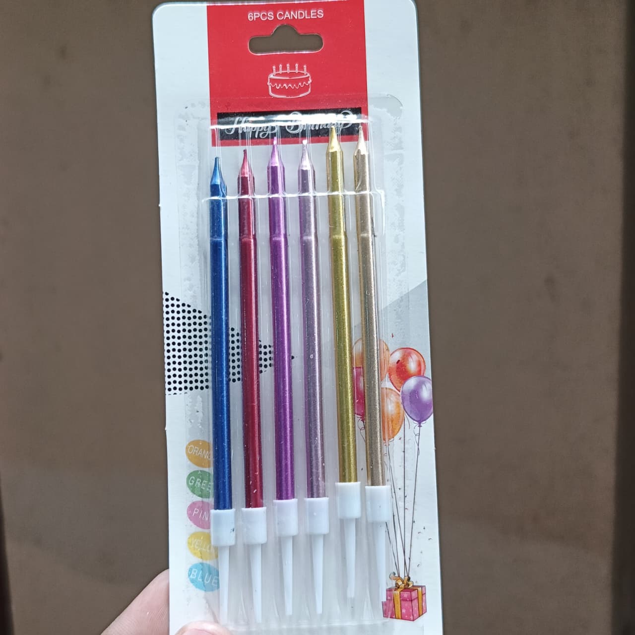 Six metallic birthday candles in blue, red, purple, gold, orange, and pink, packaged in a white and red cardboard blister pack and held by a hand against a brown background.