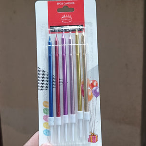 Six metallic birthday candles in blue, red, purple, gold, orange, and pink, packaged in a white and red cardboard blister pack and held by a hand against a brown background.
