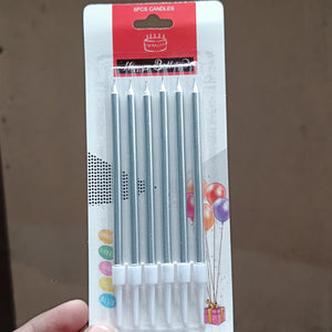 A hand holding a package of 6 silver birthday candles with white plastic bases. The packaging is white with a red header and features illustrations of balloons and a gift box.