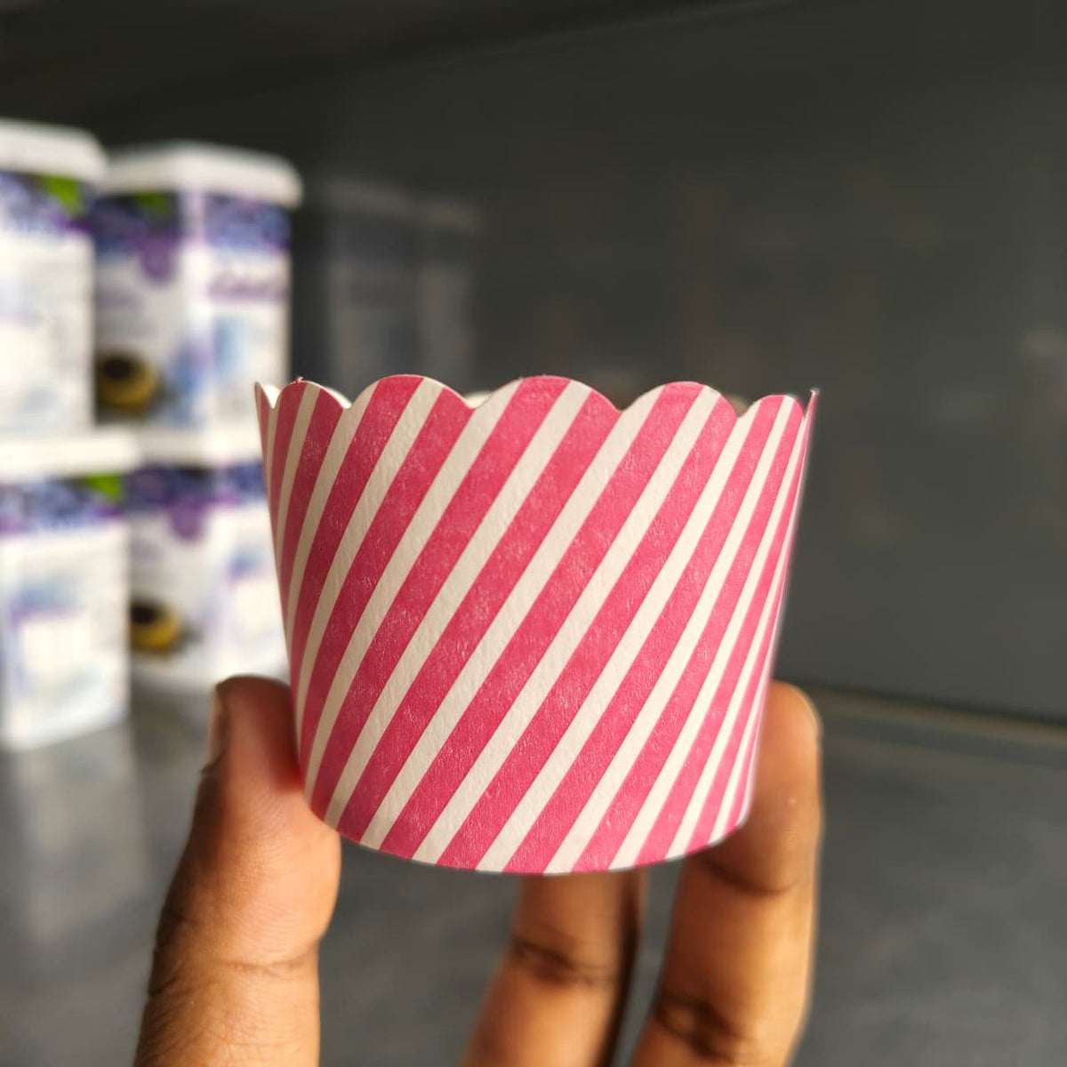A hand holding a pink and white striped scalloped cupcake liner in front of a blurred background showing other liners.