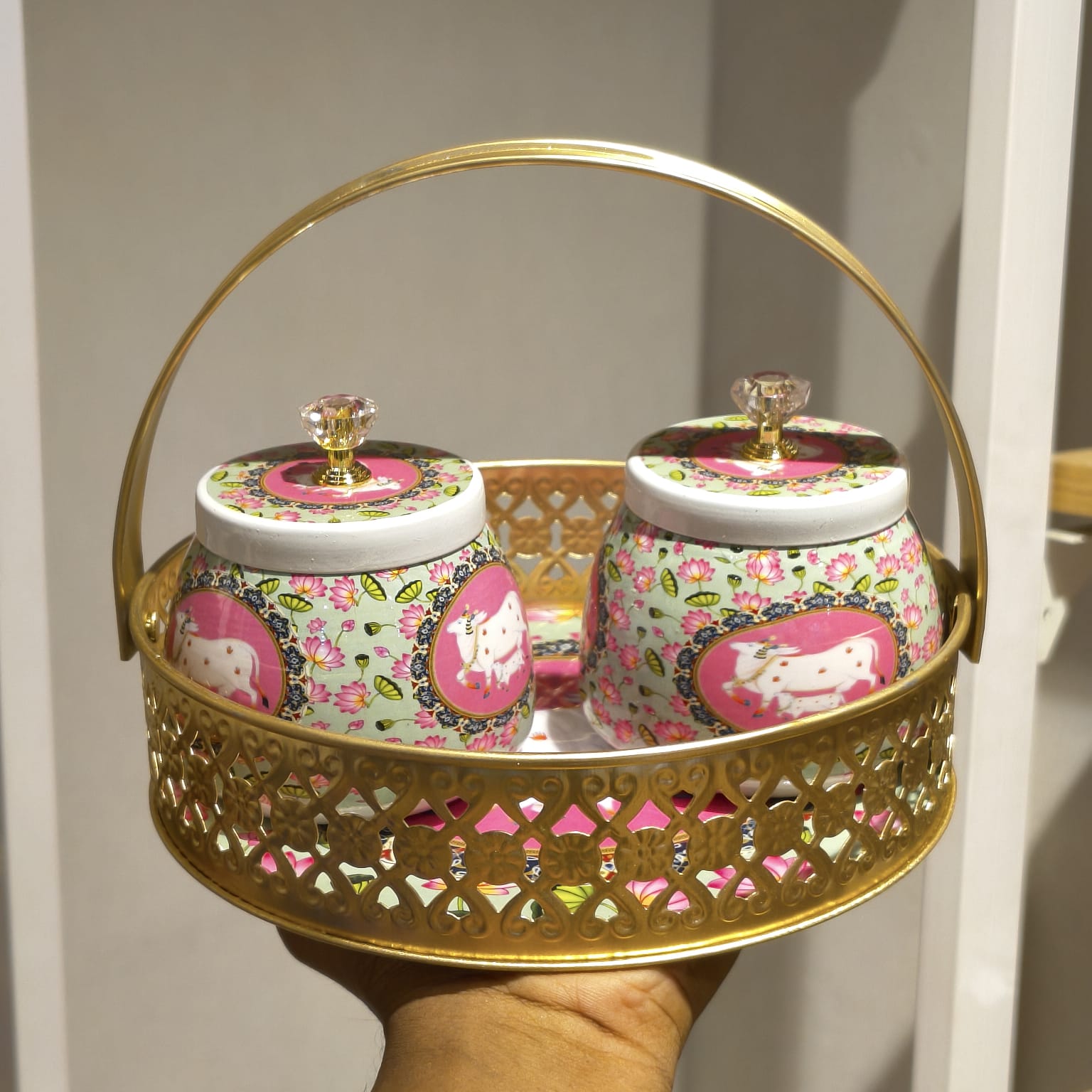 A decorative gold metal carrying basket with two matching lidded jars featuring pink floral and cow motifs, held in a hand against a neutral indoor background.