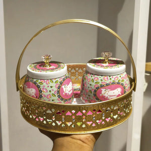 A decorative gold metal carrying basket with two matching lidded jars featuring pink floral and cow motifs, held in a hand against a neutral indoor background.