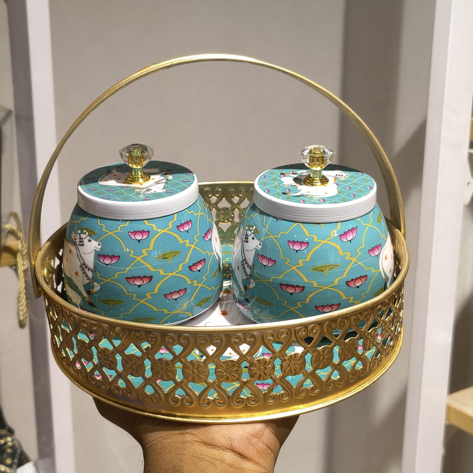 A decorative gold metal serving basket with two matching turquoise and gold patterned jars, held in a person's hand in an indoor setting.