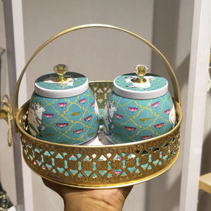 A decorative gold metal serving basket with two matching turquoise and gold patterned jars, held in a person's hand in an indoor setting.
