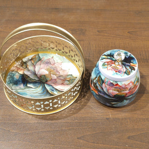 A decorative gold metal basket with a floral hand-painted interior sits next to a matching small lidded jar on a wooden tabletop. Both pieces feature colorful lotus and floral artwork in pink, blue, green, and orange tones.